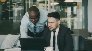 Two business partners discussing their project Bearded Caucasian businessman sitting and talking. African American colleague standing while looking at laptop computer on table.