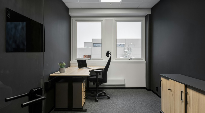 A small, empty office with a laptop open on a light-wood desk and a vacant black office chair beside two bright windows