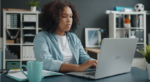 Young woman working on a laptop at a home office desk with a mug and shelving in the background